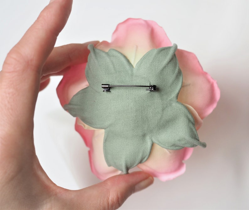 May include: A pink and white fabric flower with a green fabric backing and a metal pin on the back.