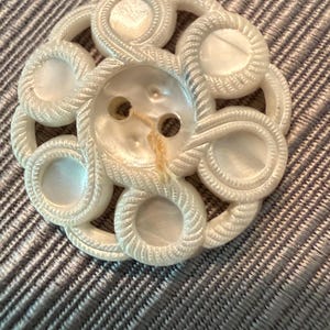 May include: A round, white, iridescent button featuring a central two-hole design surrounded by six circular cutouts. The button is decorated with a raised, textured rope-like pattern that encircles each cutout and connects to the center.