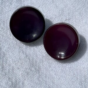 May include: Two round, dark purple buttons with a glossy finish. The buttons are set against a white, textured background, suggesting a fabric or textile. The buttons appear to be made of a smooth, solid material.