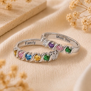 May include: Two silver-toned rings featuring a series of small, multi-colored round gemstones set in a wavy band. One ring is engraved with the word "Family" and the other is engraved with the phrase "Miss You" on the inner surface of the band.