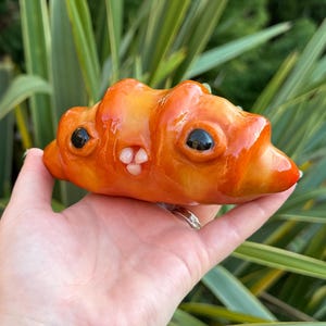 May include: An orange sculpture in the shape of a croissant, featuring a face with black eyes and a small pink nose. The artwork is held in a hand, set against a background of green leaves. The sculpture has a smiling mouth.