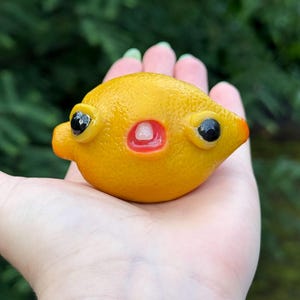 May include: A small, bright yellow, lemon-shaped figurine crafted with a textured, porous surface. It features two prominent, glossy black eyes protruding from the sides and a small, open mouth with a single white tooth visible in the centre.