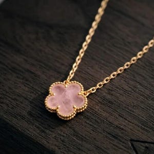 May include: A gold chain necklace featuring a five-petaled flower pendant. The pendant is crafted from a translucent light pink stone with a smooth surface, framed by a delicate gold beaded edge that matches the fine link chain.