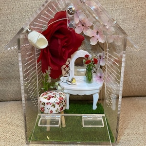May include: A miniature dollhouse scene enclosed in a clear acrylic box. The scene includes a white vanity, a floral stool, and a large red rose. Pink and white flowers and green grass add to the decor.