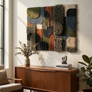 Pode incluir: Uma grande tapeçaria de parede texturizada com um design geométrico em tons de verde, laranja, azul e bege. A obra de arte é montada acima de um armário de madeira, com um vaso de eucalipto seco e uma planta em vaso em primeiro plano.