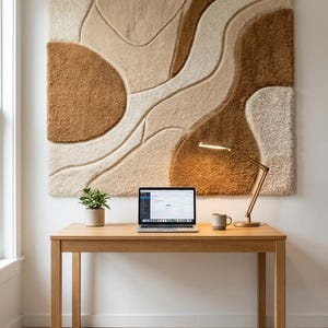 May include: A large square wall hanging featuring a tufted, textured abstract pattern in shades of cream, beige, and brown. The piece is mounted above a light-colored wooden desk that holds a laptop, a small potted plant, and a desk lamp.