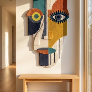 Pode incluir: Uma grande tapeçaria de parede vertical tufada com um design abstrato de rosto dividido. A peça incorpora formas geométricas em tons de amarelo mostarda, laranja queimado, azul-petróleo, azul marinho e creme, com um olho e cílios.