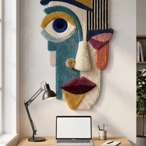 May include: Abstract face wall art in teal, cream, yellow, and burgundy. The textured artwork features a large eye, nose, and lips. The piece is mounted on a white wall above a wooden desk with a laptop and desk lamp.