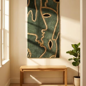 May include: A rectangular wall hanging featuring a deep green textured surface with raised gold-coloured lines forming an abstract face design. The artwork includes black almond-shaped eye and mouth details, creating a bold, linear composition.
