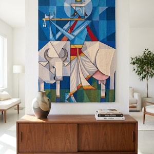 May include: A rectangular woven wall tapestry featuring a geometric blue figure playing a flute while standing beside a white cow. The artwork uses a palette of blue, white, red, and green, with sharp angular lines and a flat, stylized composition.