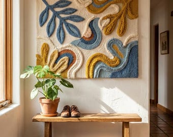 Puede incluir: Un tapiz rectangular con una superficie texturizada y copetuda que presenta formas orgánicas abstractas y motivos de hojas en tonos amarillo mostaza, azul apagado, crema y terracota, dispuestos en una composición fluida y superpuesta.
