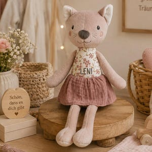 Könnte beinhalten: Ein Plüschkatzenspielzeug mit einem altrosa Körper, das ein Oberteil mit Blumenmuster und dem aufgestickten Namen LENI sowie einen passenden altrosa Rock trägt. Die Katze hat gestickte Gesichtszüge, darunter Augen, Nase und Schnurrhaare.