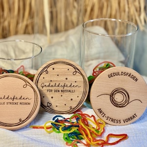 Könnte beinhalten: Drei runde Untersetzer aus hellem Holz mit deutschen Texten und dekorativen Rändern. Die Untersetzer zeigen Sätze und Designs, darunter Herzen und eine Garnrolle. Bunte Gummibänder und zwei transparente Gläser sind ebenfalls zu sehen.