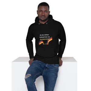 Puede incluir: Sudadera con capucha negra con el texto alemán "Ab der dritten Halbzeit bin ich Bundestrainer" y un gráfico con los colores de la bandera alemana. La persona lleva vaqueros azules.