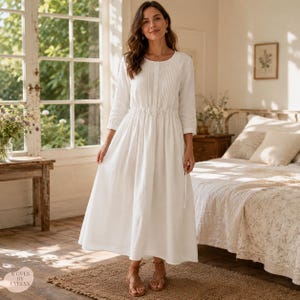 May include: A white midi-length dress with three-quarter sleeves, a round neckline, and vertical pleat detailing on the bodice. The garment features a cinched waist with a drawstring tie and a full, flowing skirt. The text Woven by Evelyn is visible.