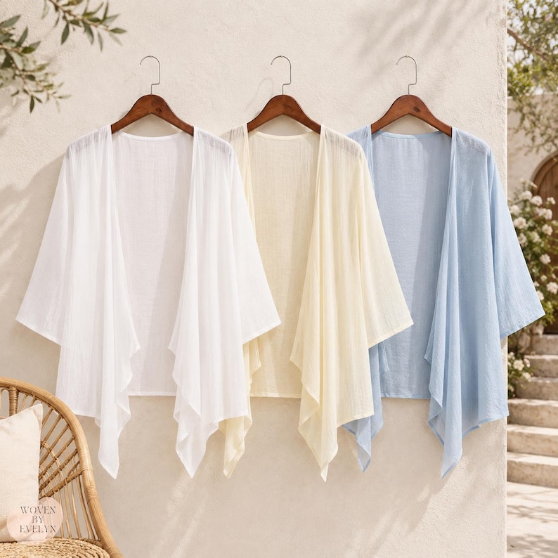Puede incluir: Tres c&aacute;rdigans abiertos colgados en perchas de madera, presentados en blanco, amarillo p&aacute;lido y azul claro. Cada prenda presenta una silueta holgada y drapeada con mangas anchas de tres cuartos y un tejido ligero y texturizado.