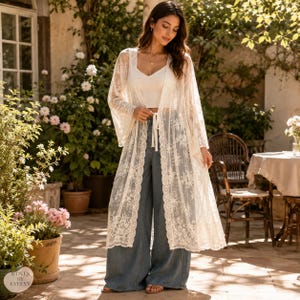 May include: A long, cream-colored lace duster cardigan featuring wide sleeves and a delicate floral pattern, worn over a simple white camisole and loose-fitting blue denim jeans. The garment has an open front with a tie closure at the waist.