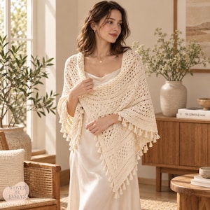 May include: A cream-colored triangular crochet shawl featuring an intricate openwork pattern and a decorative fringe of small tassels along the edges. The shawl is draped over a simple, sleeveless cream-colored dress with a smooth, satin-like finish.