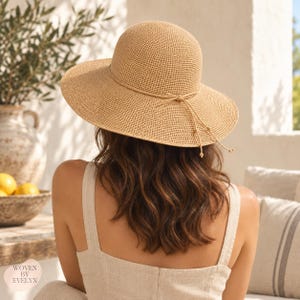 May include: A light tan woven sun hat with a rounded crown and a wide brim. A thin matching cord wraps around the base of the crown, tied in a small bow at the front with two dangling ends. The hat features a fine, uniform texture throughout.