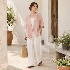 Puede incluir: Una chaqueta tipo kimono de color rosa empolvado abierta por delante con mangas tres cuartos, sobre una camiseta de tirantes blanca y pantalones blancos de pernera ancha. Se combina con sandalias marrones y un bolso de paja. El texto Woven by Evelyn es visible.