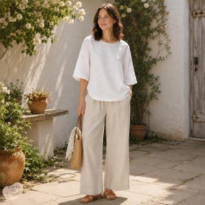 May include: A white short-sleeved top with a round neckline paired with loose-fitting beige trousers. The outfit is completed with brown sandals and a woven straw handbag featuring a patterned fabric accessory attached to the handle.