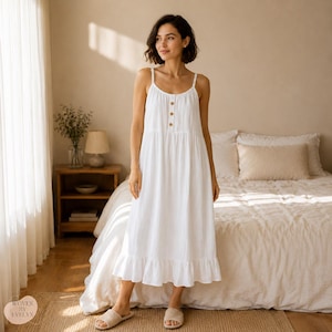 May include: A white midi-length dress featuring thin spaghetti straps, a scoop neckline, and a vertical row of three brown buttons on the bodice. The garment has a textured, crinkled fabric finish and a ruffled hemline at the bottom of the skirt.