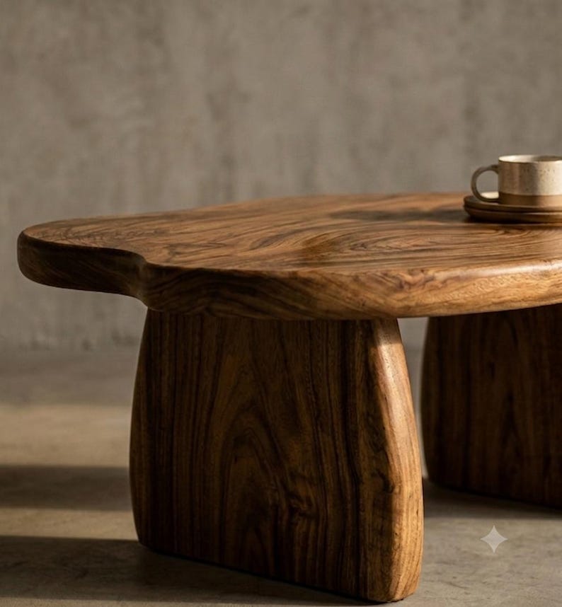 May include: A low-profile coffee table crafted from solid wood with a natural brown finish and visible grain patterns. The table features an organic, irregular tabletop shape supported by two thick, curved slab legs. A ceramic mug sits on the surface.