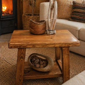 May include: A square wooden coffee table with a lower shelf, holding a decorative wooden bowl filled with small wooden objects. The table is made of light brown wood and has a natural, rustic appearance. A wooden sculpture and a wooden bowl sit on top.