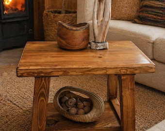 May include: A square wooden coffee table with a lower shelf, holding a decorative wooden bowl filled with small wooden objects. The table is made of light brown wood and has a natural, rustic appearance. A wooden sculpture and a wooden bowl sit on top.
