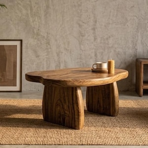 May include: A low-profile coffee table crafted from solid wood with a natural grain pattern, featuring an irregular, organic-shaped tabletop supported by two thick, curved slab legs. A ceramic mug and a textured pillar candle sit on top.