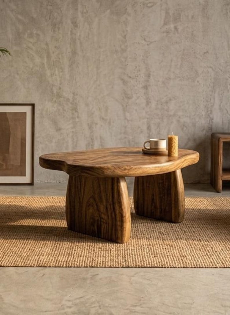 May include: A low-profile coffee table crafted from solid wood with a natural grain pattern, featuring an irregular, organic-shaped tabletop supported by two thick, curved slab legs. A ceramic mug and a textured pillar candle sit on top.