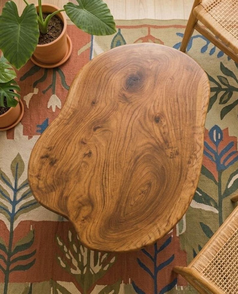 May include: A coffee table with an irregular, organic shape crafted from natural wood. The surface displays a prominent grain pattern with visible knots and swirls in warm brown tones. The edges are smooth and rounded, following the natural contour of the wood slab, creating a distinct, non-geometric silhouette that serves as a central piece of furniture.