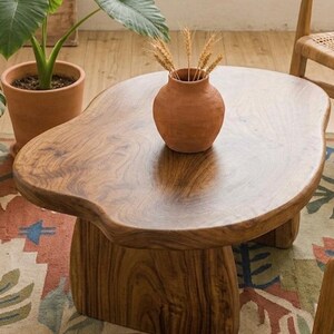 May include: A low coffee table crafted from natural wood with an irregular, organic-shaped top and a thick, solid base. A small, round, terracotta-colored ceramic vase containing several dried wheat stalks sits on the center of the table surface.