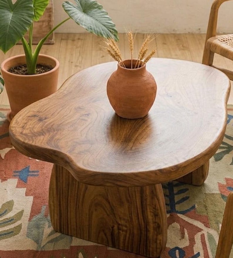 May include: A low coffee table crafted from natural wood with an irregular, organic-shaped top and a thick, solid base. A small, round, terracotta-colored ceramic vase containing several dried wheat stalks sits on the center of the table surface.