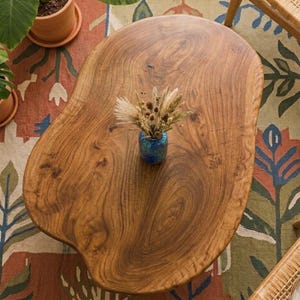 May include: A natural-edged, organic-shaped wooden coffee table with a visible wood grain pattern. Centered on the table is a small, round, blue ceramic vase containing a dried floral arrangement with beige and brown grasses and seed pods.