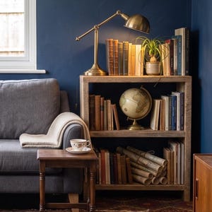 May include: A cozy living room scene featuring a wooden bookshelf filled with books, a globe, and a small potted plant. A brass desk lamp illuminates the top shelf. A gray sofa with a blanket and a small wooden side table with a teacup complete the scene.