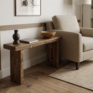 May include: A rustic wooden side table with a dark stain, featuring a rectangular top and thick, sturdy legs. A dark vase and a gray notebook rest on the table, alongside a wooden bowl. The table is next to a beige armchair, creating a cozy living room scene.
