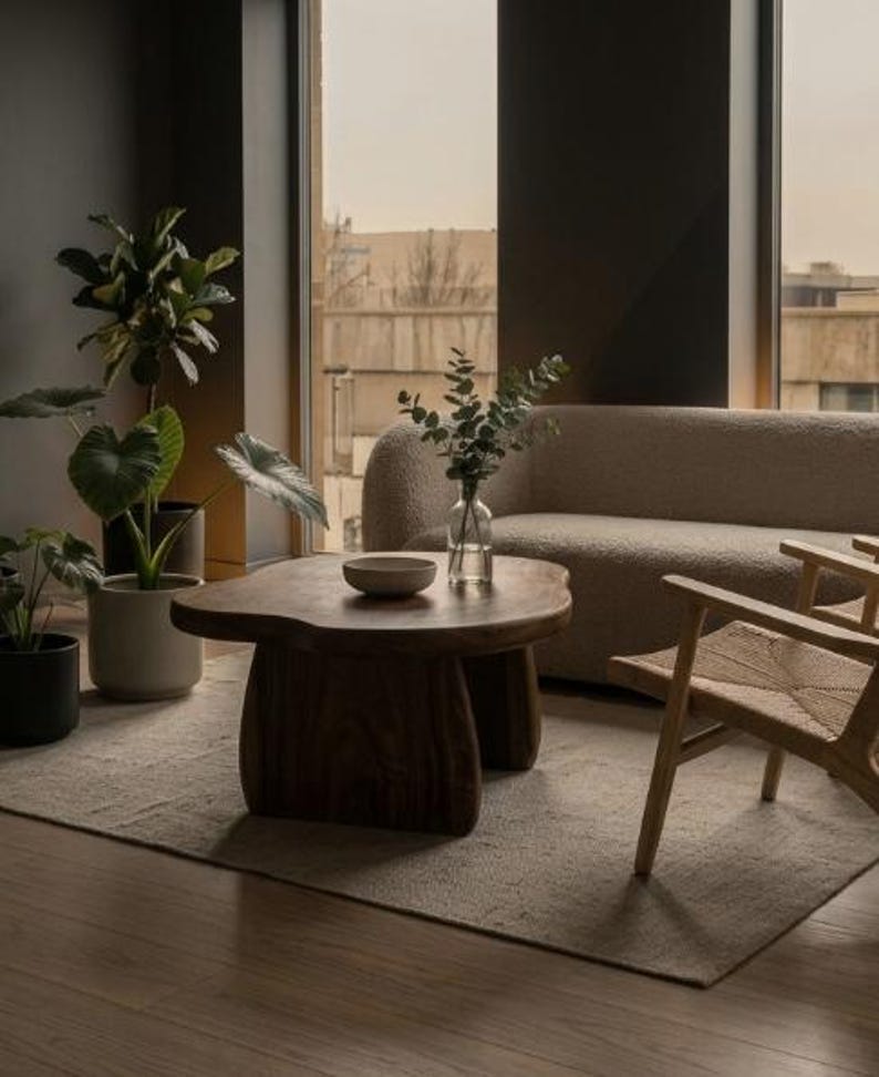May include: A dark wood coffee table with an organic, irregular shape and thick, curved legs. On the table sits a small, shallow bowl and a clear glass vase containing green leafy stems. The table is positioned in front of a beige textured sofa.