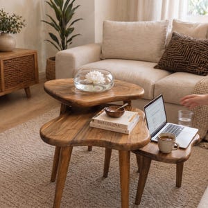 May include: Three wooden, organically shaped side tables of varying sizes. A clear glass bowl with a white flower sits on the top table. A laptop, mug, and book are on the lower tables. The tables are in a living room setting.