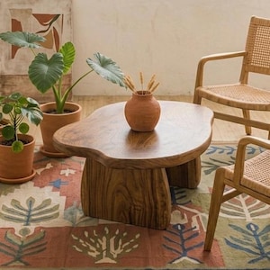 May include: A low, organic-shaped wooden coffee table with a smooth, dark brown grain surface and two thick, curved slab legs. The table is positioned in a room with a patterned rug, two potted green plants, and two light-colored wooden chairs.
