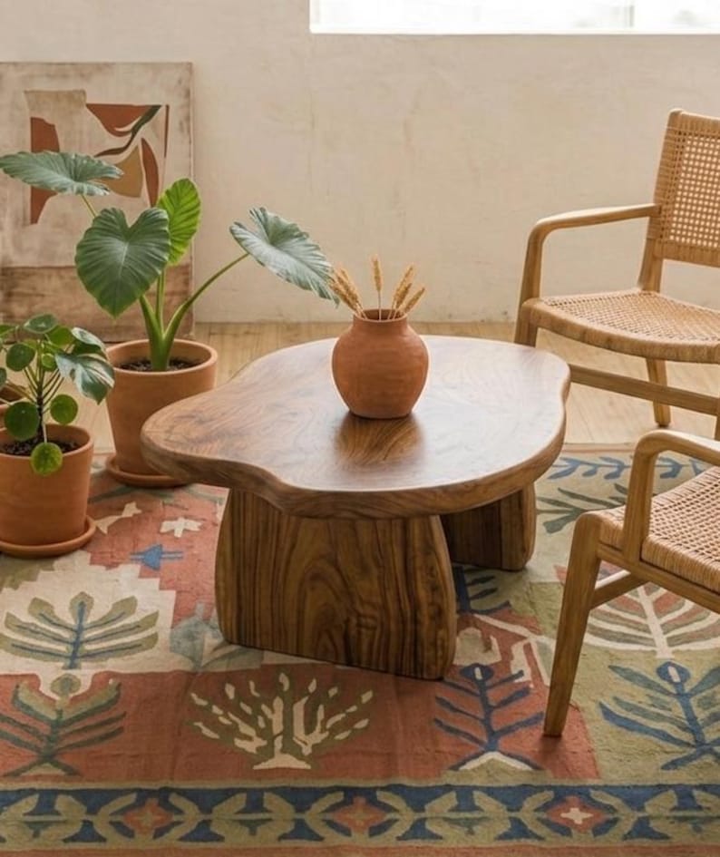 May include: A low, organic-shaped wooden coffee table with a smooth, dark brown grain surface and two thick, curved slab legs. The table is positioned in a room with a patterned rug, two potted green plants, and two light-colored wooden chairs.