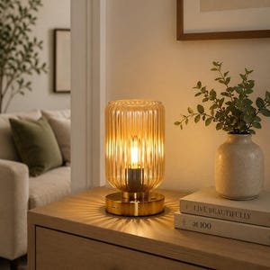 May include: A decorative amber glass table lamp with a ribbed design, resting on a gold-colored base. The lamp is illuminated, casting a warm glow. The lamp is on a wooden nightstand with books and a vase.