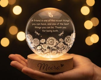 May include: A clear glass sphere featuring an etched sunflower field, a butterfly, and two dragonflies. The text reads: A friend is one of the nicest things you can have, and one of the best things you can be. Thank you for being both. It sits on a light wood base with the name Meera engraved on the front.