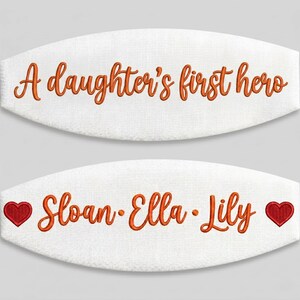 May include: Two white oval-shaped fabric patches featuring orange embroidered text. The top patch reads A daughter's first hero. The bottom patch displays the names Sloan, Ella, and Lily in orange, flanked by two small red embroidered hearts.