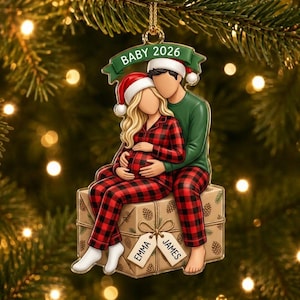May include: A Christmas ornament featuring two figures sitting on a gift box. One figure wears red and black plaid pyjamas and a Santa hat, the other wears a green shirt and Santa hat. A green banner reads BABY 2026. Gift tags are labelled EMMA and JAMES.