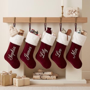 May include: Five deep red velvet Christmas stockings with white faux fur cuffs, each personalised with white embroidered names: Sophia, James, Lily, Leo, and Mia. The stockings hang from a wooden mantel, filled with various small gifts and toys.