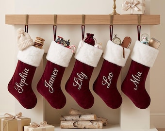May include: Five deep red velvet Christmas stockings with white faux fur cuffs, each personalized with white embroidered names: Sophia, James, Lily, Leo, and Mia. The stockings hang from a wooden mantel, filled with various small gifts and toys.