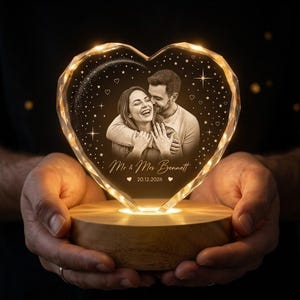 May include: A heart-shaped glass plaque with an illuminated edge, featuring an engraved portrait of a couple, the text "Mr & Mrs Bennett", and the date "20.12.2026". The plaque is set on a round wooden base and decorated with stars and hearts.