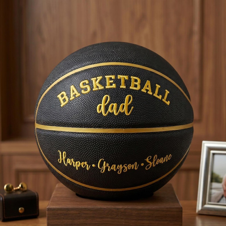 May include: A black basketball with gold textured lines and gold lettering. The text reads "BASKETBALL dad" in a mix of block and script fonts, with the names "Harper", "Grayson", and "Sloane" printed in gold script below the center line.