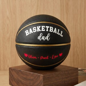 May include: A black basketball featuring gold lines and white text that reads "BASKETBALL dad". Below the text, the names "Oliver", "Pearl", and "Ezra" are printed in red script, flanked by two small red heart icons on the textured surface.